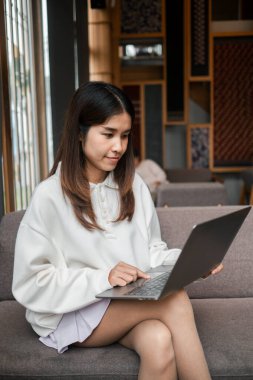Gündelik kıyafetli genç bir kadın modern bir oturma odasında dizüstü bilgisayar ve doğal ışıklandırmayla koltukta oturur..