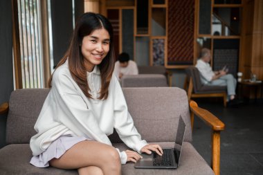 Genç bir kadın, sıcak, ahşap bir iç mekanı olan şık bir kafede dizüstü bilgisayarıyla çalışırken gülümsüyor..