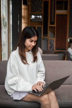 Beyaz kazaklı genç bir kadın, modern bir odada, doğal ışığı olan bir dizüstü bilgisayar kullanarak koltukta oturuyor..