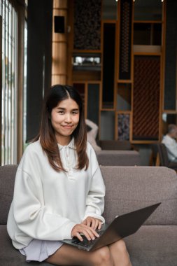 Genç bir kadın, şık dekorlu ve doğal ışıklandırmalı modern bir kafede dizüstü bilgisayarıyla çalışırken gülümsüyor..