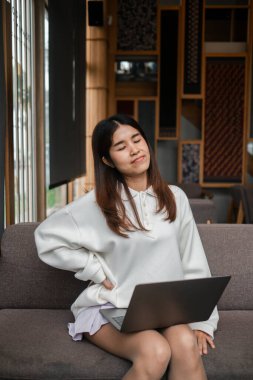 Genç bir kadın bir kanepede dizüstü bilgisayarla rahat bir şekilde oturur ve modern bir oturma odasında huzurlu bir anın tadını çıkarır..