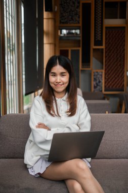 Ahşap dekor ve doğal ışıkla çevrili şık bir kafede dizüstü bilgisayarla kanepede oturan gülümseyen genç kadın..