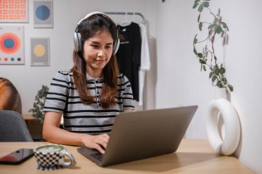 Kadın dizüstü bilgisayara odaklanmış, kulaklık takıyor, bitkileri ve modern dekorları olan şık bir ev ofisinde çalışıyor..