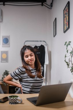 Çizgili tişörtlü genç bir kadın, ofiste çalışırken sırt ağrısı çekiyor. Modern dekorasyonla çevrili..