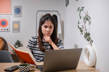 Evde işine konsantre olan genç bir kadın, şık, minimalist bir çalışma alanında dizüstü bilgisayar ve defter kullanıyor..