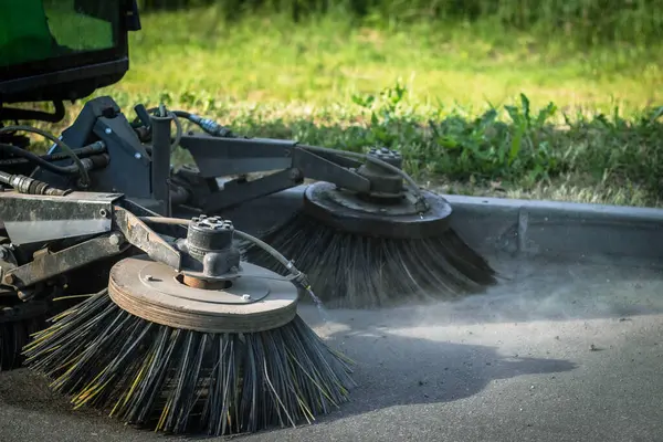 İki dönen fırçayla asfalt kaldırımı temizleyen bir sokak süpürgesinin yakın plan fotoğrafı. Süpürge yeşil ve ön planda büyük bir lastiği var. Fırçalar sert kıllardan yapılmıştır ve enkazı temizlemek için dönüyorlar. Süpürge park etti.