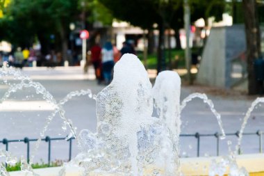 Parktaki çeşmeden zarif bir şekilde su sıçrıyor. Gününün tadını çıkaran insanlarla dolu..
