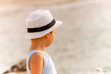 Suyun kenarında duran, hasır şapkalı ve mavi gömlekli genç bir çocuk güneşli bir günde sakin denizi seyrediyor..