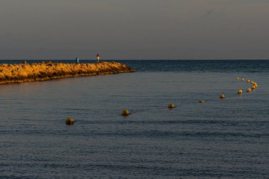 Bir birey gün batımında kayalık bir iskelede duruyor, yüzen şamandıralarla sakin denize bakıyor..