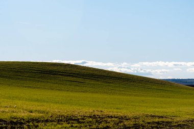Geniş yeşil alan yavaşça yükseliyor, parlak mavi gökyüzü ile sakin bir kırsal ortam sergiliyor..