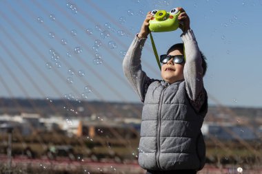 Parkta güneşli bir günde genç bir çocuk gülümsüyor ve canlı bir kamerayla kabarcıkları yakalıyor..