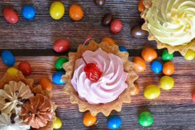 Renkli turtalar, protein kremalı kekler, ahşap masada çok renkli Dragee şekerler. Çikolatalı, kirazlı ve muzlu lezzetli bir tatlı, kısa hamurlu pasta. Parti yemeği. Üst görünüm.