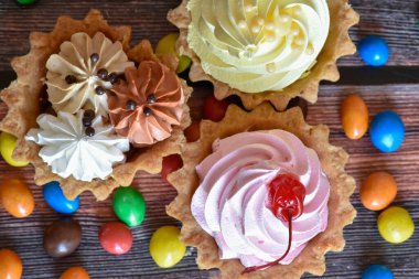 Renkli turtalar, protein kremalı kekler, ahşap masada çok renkli Dragee şekerler. Çikolatalı, muzlu ve kirazlı lezzetli tatlı, kıtır ekmek. Parti yemeği. Üst görünüm.