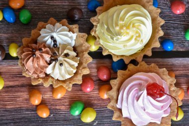 Renkli turtalar, protein kremalı kekler, ahşap masada çok renkli Dragee şekerler. Çikolatalı, muzlu ve kirazlı lezzetli tatlı, kıtır ekmek. Parti yemeği. Üst görünüm.