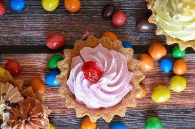 Renkli turtalar, protein kremalı kekler, ahşap masada çok renkli Dragee şekerler. Çikolatalı, kirazlı ve muzlu lezzetli bir tatlı, kısa hamurlu pasta. Parti yemeği. Üst görünüm.