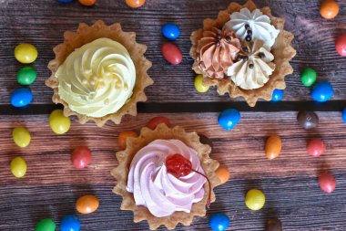 Renkli turtalar, protein kremalı kekler, ahşap masada çok renkli Dragee şekerler. Muz, çikolata ve kiraz aromalı lezzetli tatlı, kısa hamurlu pasta. Parti yemeği. Üst görünüm.