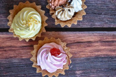 Ahşap masa arkasında renkli protein kremi olan kekler. Muz, çikolata ve kiraz aromalı lezzetli tartlar, kısa hamurlu pastalar. Üst görünüm.