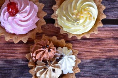 Ahşap masa arkasında renkli protein kremi olan kekler. Kiraz, muz ve çikolata aromalı lezzetli tartlar, kısa hamurlu pastalar. Üst görünüm.