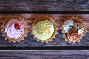 Ahşap masa arkasında renkli protein kremi olan kekler. Kiraz, muz ve çikolata aromalı lezzetli tartlar, kısa hamurlu pastalar. Üst görünüm.