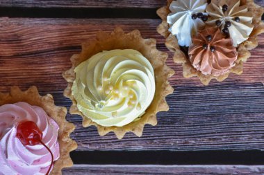 Ahşap masa arkasında renkli protein kremi olan kekler. Kiraz, muz ve çikolata aromalı lezzetli tartlar, kısa hamurlu pastalar. Üst görünüm.