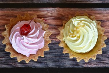 Ahşap masa arkasında pembe ve sarı protein kremalı turtalar. Kiraz ve muz aromalı lezzetli bir tatlı, kısa hamurlu pasta. Üst görünüm.