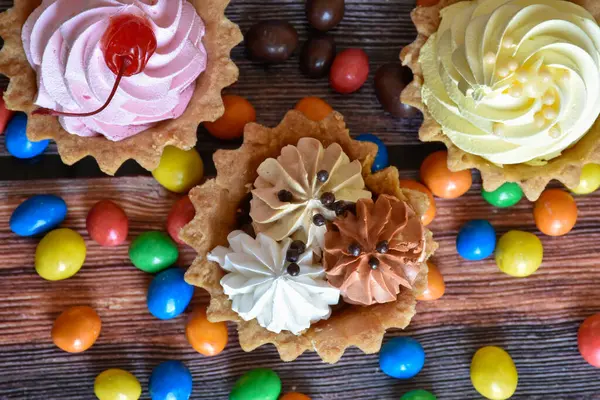 Renkli turtalar, protein kremalı kekler, ahşap masada çok renkli Dragee şekerler. Kirazlı, çikolatalı ve muzlu lezzetli bir tatlı, kısa hamurlu pasta. Parti yemeği. Üst görünüm.