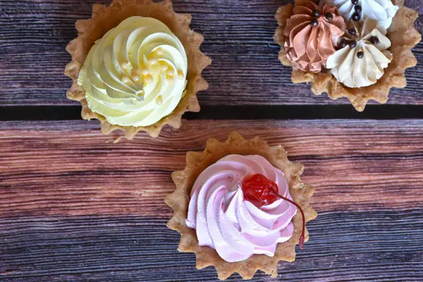 Ahşap masa arkasında renkli protein kremi olan kekler. Muz, çikolata ve kiraz aromalı lezzetli tartlar, kısa hamurlu pastalar. Üst görünüm.