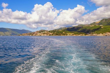 Yaz tatili konsepti. Akdeniz manzarası. Karadağ, eğlence gemisinden Kotor Körfezi manzarası. Herceg Novi şehri uzakta. Teknenin arkasında dalgalı su izleri var. Güzel Kümülüs bulutları