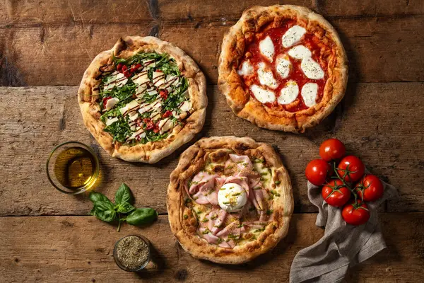 Üzerinde çeşitli malzemelerle üç farklı pizzanın kırsal ahşap bir yüzey üzerinde görüntüsü var..