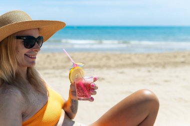Parlak bikinili seksi bir sarışın elinde deniz kenarında bir kokteyl ile güneşleniyor. Güzel bir kız yaz tatilinin tadını çıkarıyor. Pahalı bir tatil köyünde, kaliteli bir fotoğrafta.