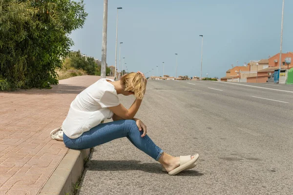 stock image a girl sits on the pavement in jeans and a T-shirt, turned her back to the camera, looks at the road where cars drive. High quality photo