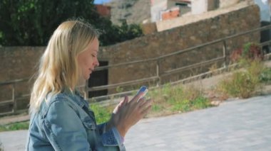 Güzel saçlı mutlu kız sokakta bir bankta otururken telefonuna selfie çekiyor. Yüksek kalite 4k görüntü