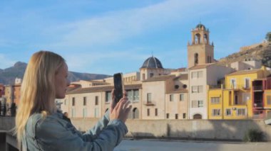 Modern sarışın bir kadın binaların yanında dikiliyor ve manzaranın tadını çıkarmak için kamerasını akıllı telefonuna çeviriyor. Yüksek kalite 4k görüntü
