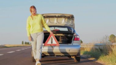 Sarı saçlı, sarı kazaklı ve kot pantolonlu bir kız otoyola kırık bir arabanın yanına acil dur işareti koyuyor. Araba yolda arıza yaptı. Yüksek kalite 4k görüntü