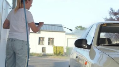 a cheerful girl in a white t-shirt with blond hair tied in a ponytail washes a gray car with water outdoors.The concept of work. High quality photo