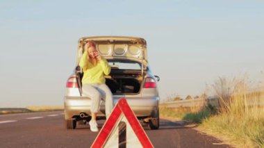 Sarı kazaklı ve Avrupalı görünümlü bir kız bagajda oturuyor ve telefonda duygusal olarak konuşuyor, yolda bir olay, ön planda bir uyarı üçgeni. Yüksek kalite 4K görüntü.