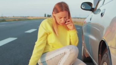 a girl in jeans of European appearance sits near the rear wheel of a punctured one, speaks on the phone how to make a wheel. With a surprised and incomprehensible face what to do High quality photo