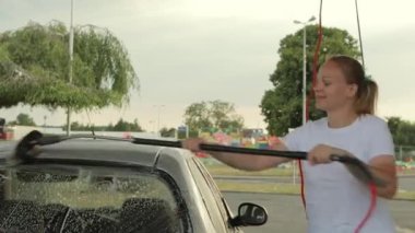 Beyaz tişörtlü, kotlu, sarı saçlı bir kız oto yıkamada araba yıkıyor.