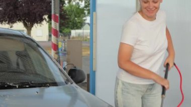 Beyaz tişörtlü, kotlu, sarı saçlı bir kız oto yıkamada araba yıkıyor.
