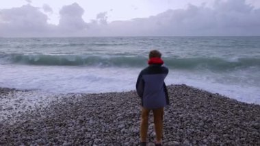 Ceketli neşeli genç çocuk okyanus kıyısında çakıl taşlarının üzerinde duruyor, sırtı kameraya dönük, dalgalarla eğleniyor, mutlu bir çocukluk geçiriyor. Yüksek kalite 4k görüntü