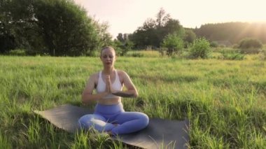 Genç bir kadın, yaz günbatımında yeşil çimlerin arkasında yoga yapıyor. Temiz hava egzersizi. Sağlık hizmeti, özgünlük, denge ve sakinlik duygusu. Yüksek kaliteli FullHD görüntüler
