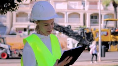 İş sırasında inşaat sahasında duran, kasketli ve çelik yelekli Avrupalı genç bir kadın, dokümanları okuyor. Yüksek kaliteli FullHD görüntüler