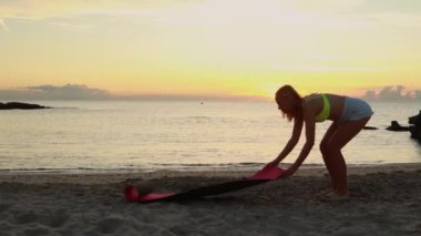 İnce bir kadın dalgaların yanındaki deniz kıyısında yoga yapıyor. Şafakta güneş ufukta beliriyor. Yüksek kalite 4k görüntü
