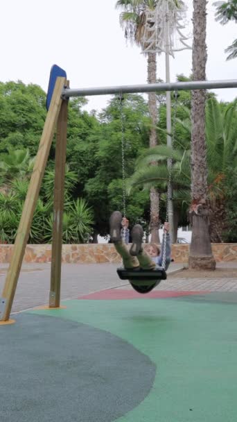 垂直ビデオ 晴れた日の公園でスイングする少年 スイングで遊び場にいる子供 低アングルショット 遊び場で白人の子供 高品質の映像 — ストック動画