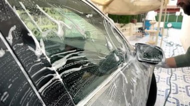 Bir adam evin önünde köpükle gri bir arabayı yıkar, evde araba yıkama kavramı. Yüksek kaliteli FullHD görüntüler