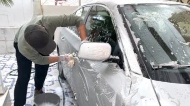 Siyah şapkalı ve günlük kıyafetli bir adam evin önünde köpüklü gri bir arabayı yıkıyor. Evde araba yıkama kavramı. Yüksek kaliteli FullHD görüntüler