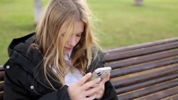 かわいい美しい女の子は 市の公共公園のベンチに一人で座り スマートフォンでビデオゲームをしています 子供は喜んで笑い 携帯電話の垂直モードを使用しています 高品質の4K映像 — ストック動画