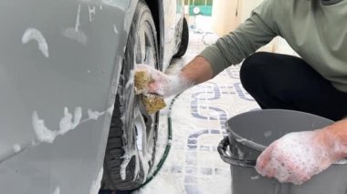 Elinde süngerle bir adamın eli ve yakınlarda duran bir araba için gri bir kova deterjan evin bahçesinde arabaların alaşımlı tekerlerini yıkıyor. Yüksek kaliteli FullHD görüntüler