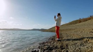 Bir kadın kayalık bir sahilde ve akıllı telefonuyla video çekiyor. Yürüyüş konsepti. Sıradan giyinmiş bir kız. Yüksek kalite 4k görüntü