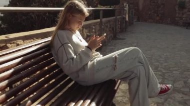 Güzel sarışın bir genç, şehir parkında bir bankta oturuyor, cep telefonu uygulamalı bir telefon kullanıyor, sosyal ağları kullanıyor, haberleri kontrol ediyor. Kamera, kızın yüksek kaliteli 4K görüntülerine odaklanıyor.